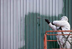 Peintre professionnel à Granby portant un équipement de protection complet sur une nacelle. Il peinture le revêtement d'un bâtiment en métal.
